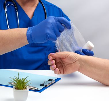 doctor sits at a table in blue uniform and latex gloves, specialist bandages his hand with a white bandage to the patient, receiving patients in the office