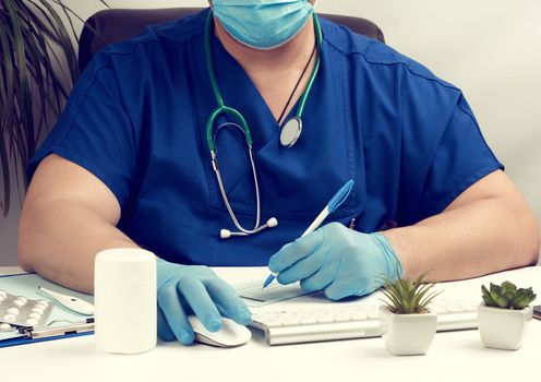 male doctor sitting at a white table in blue uniform and latex gloves, specialist writes a prescription for a pharmacy, medical office for receiving patients