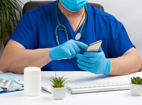 doctor in a blue uniform sits at a desk and holds in his hand a smartphone, concept of distance medicine and consultations