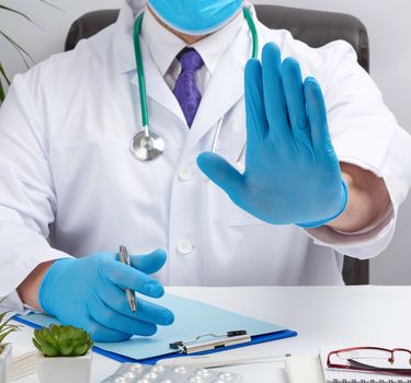 doctor in a white medical coat sits at a table in a brown leather chair and shows with his palm a gesture of stop, calm down and not panic