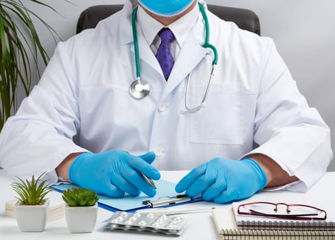 doctor in a white uniform sits at a white table and writes in a paper notebook, wearing sterile gloves on his hands, concept of receiving patients in the office