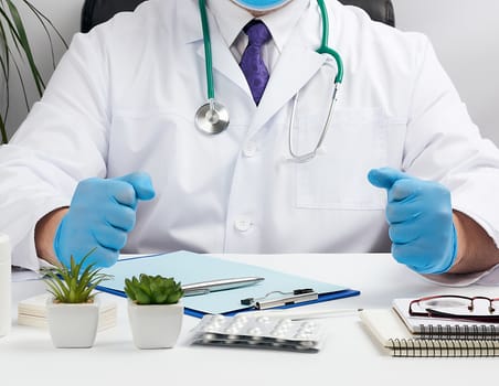 doctor in a white coat and blue latex gloves sits at a white work table in his office, concept of patient reception in the clinic, focused pose