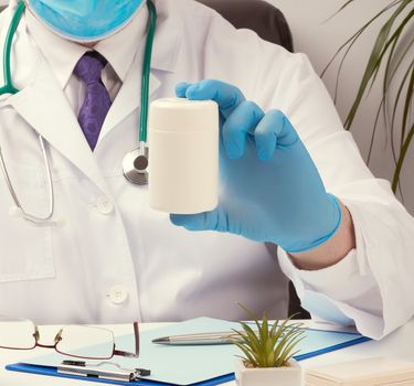 male doctor in a white coat and blue latex gloves sits at a white desk in his office and holds a plastic white jar of pills