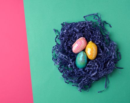 decorative Easter eggs from sequins lie in a nest of strips of paper on a colored background, top view
