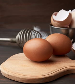 brown whole chicken eggs on a wooden board, behind a whisk for whipping and a shell
