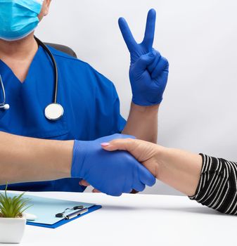 doctor in a blue uniform sits at a desk in an armchair and shakes hands with a female patient, concept of gratitude, greetings, another hand shows victory gesture