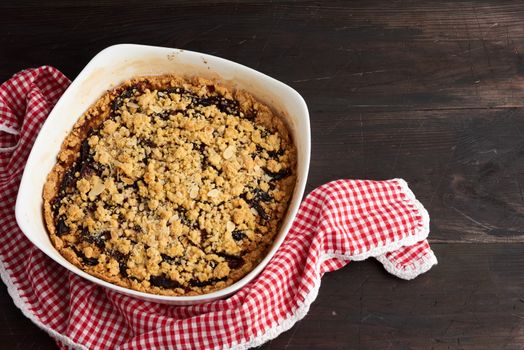 baked crumble pie with blue plum in a white ceramic plate on a wooden table, tasty dessert, top view