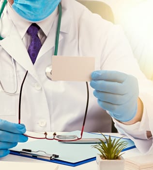 doctor in a white medical coat sits at a table in a brown leather chair and holds a rectangular white blank business card in his hands, medical office