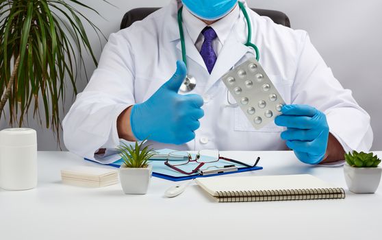 male doctor in a white coat and blue latex gloves sits at a white desk in his office and shows a pack of pills in a blister, with his other hand shows a gesture like