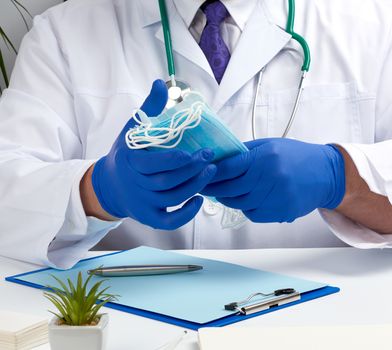 doctor in a white coat and blue latex gloves sits at a white desk and shows a stack of disposable medical masks, protective agent against viruses and stopping the spread of the epidemic