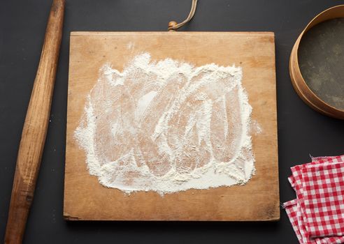 white wheat flour scattered on a black table and a wooden rolling pin, round sieve on a black table, top view