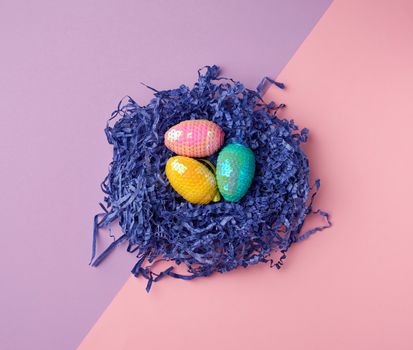 decorative Easter eggs from sequins lie in a nest of strips of paper on a colored background, top view