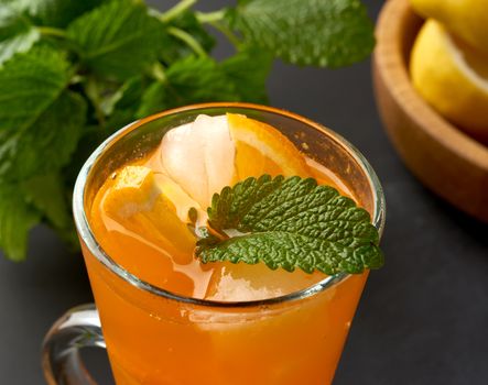 transparent large glass with a handle filled with lemonade with slices of orange and lemon, behind a bunch of green mint, black background