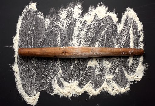 white wheat flour scattered on a black table and very old brown wooden rolling pin on a black table, top view