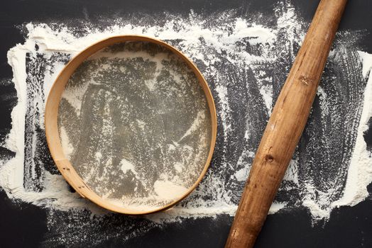 white wheat flour scattered on a black table and very old brown wooden rolling pin, round sieve, top view