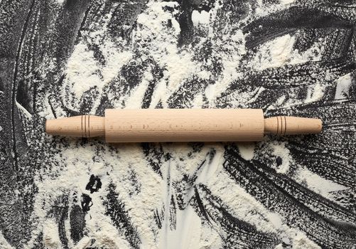 white wheat flour scattered on a black table and a wooden rolling pin on a black table, top view