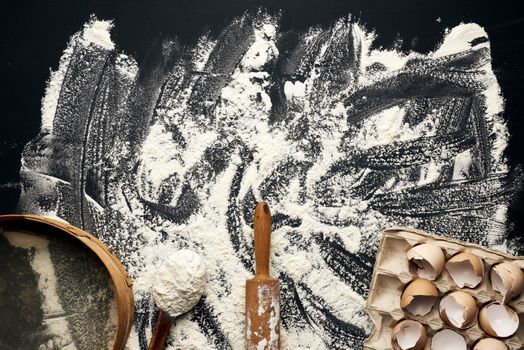 sprinkled white wheat flour, wooden rolling pin, round sieve and chicken eggshell in a paper tray, top view, kitchen backdrop