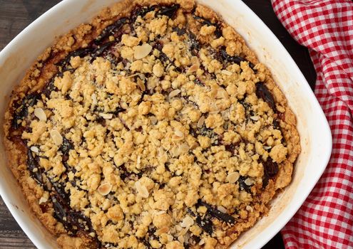 baked crumble pie with blue plum in a white ceramic plate on a wooden table, tasty dessert, top view