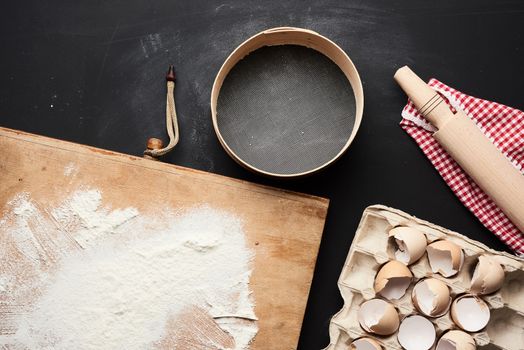 wooden round sieve, white wheat flour, tray of chicken eggs in a tray and rolling pin on a black table, ingredients for kneading dough, top view