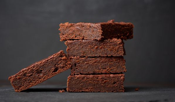 stack of square baked pieces of brownie chocolate cake on a black background, delicious dessert