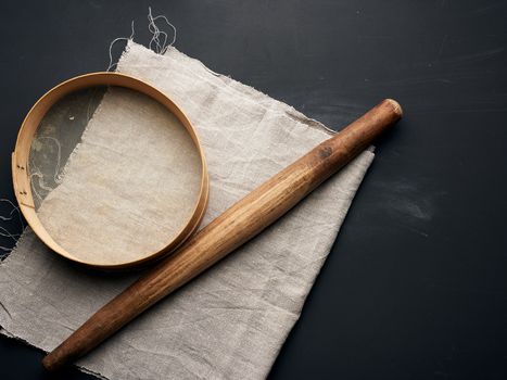 vintage wooden rolling pin and a round sieve lie on a gray linen napkin, black table, top view