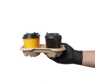 hand in a black latex glove holds a paper tray with disposable cardboard cups and plastic covers on a white background, safe and contactless delivery of on-line orders during epidemics