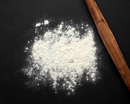 white wheat flour scattered on a black table and very old brown wooden rolling pin on a black table, top view