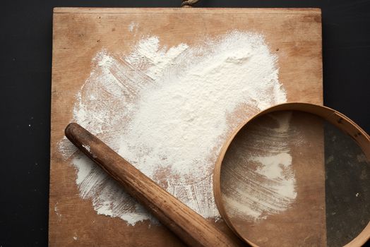 white wheat flour scattered on a black table and a wooden rolling pin, round sieve on a black table, top view