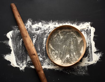 white wheat flour scattered on a black table and very old brown wooden rolling pin, round sieve, top view