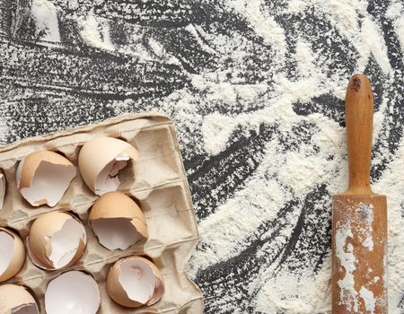 sprinkled white wheat flour, wooden rolling pin and chicken eggshell in a paper tray, top view, kitchen backdrop