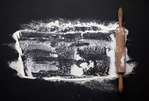 white wheat flour scattered on a black table and a wooden rolling pin on a black table, top view, place for an inscription