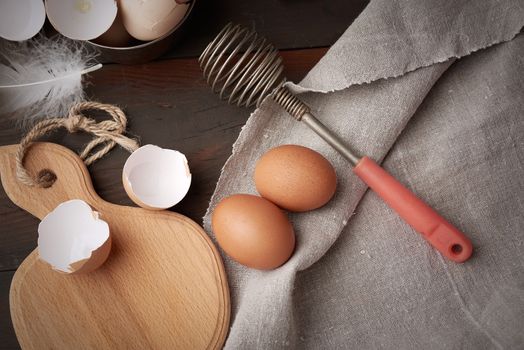 brown raw chicken eggs, shells, cutting board and hand-held whisk for protein on a wooden table, top view