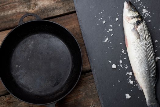 fresh whole sea bass fish on a black board, next to it is an empty round black frying pan on a wooden table, top view