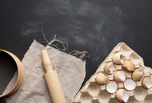 wooden round sieve, tray of chicken eggs and rolling pin on a black table, ingredients for kneading dough, top view, copy space