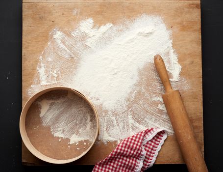 white wheat flour scattered on a black table and a wooden rolling pin, round sieve on a black table, top view