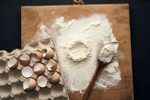 sprinkled white wheat flour, wooden rolling pin, round sieve and chicken eggshell in a paper tray, top view, kitchen backdrop