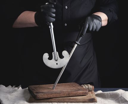 male cook in black uniform and black latex gloves holds a large sharp vintage knife for meat and vegetables and a metal sharpener for knives, black background