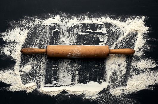 white wheat flour scattered on a black table and a wooden rolling pin on a black table, top view, place for an inscription