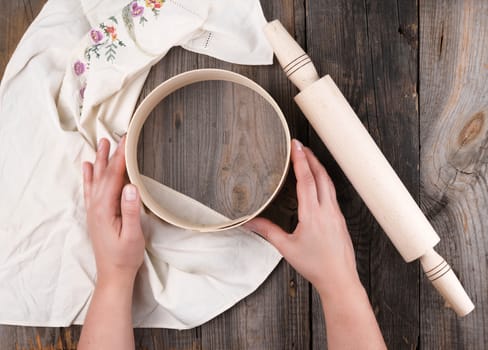 female hands holding a round wooden sieve for flour, next to an wooden rolling pin on a gray table