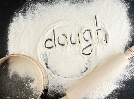 sprinkled white wheat flour on a black table and the inscription dough, next to a wooden round sieve and rolling pin, top view