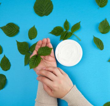 female hands of a young girl with smooth skin and round jar of cream, blue background with green raspberry leaves, concept of spa treatments for skin
