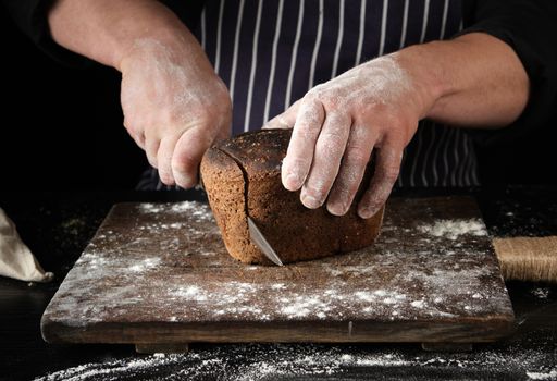 chef in black uniform holds a kitchen knife in his hand and cuts off pieces of bread from a baked brown rye flour loaf on a wooden board
