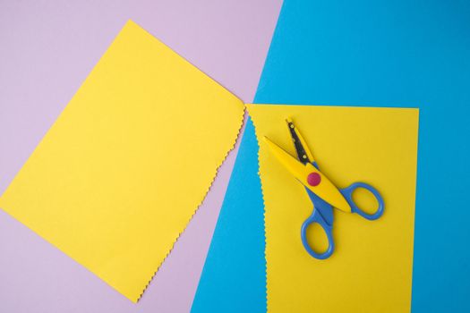 abstract background of colored paper, yellow stripes with a zigzag end and plastic scissors, top view