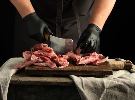 chef in black latex gloves holds a big knife and cuts into pieces raw rabbit meat on a brown wooden cutting board, cooking on a dark background