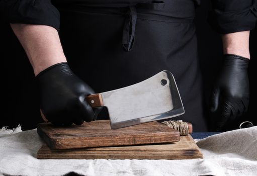 male cook in black uniform and black latex gloves holds a large sharp meat knife over a cutting board, black background