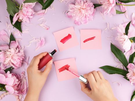 female hands with smooth fair skin keep liquid red lipstick in a tube on a lilac background with pink peonies, top view