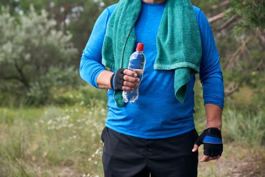 adult athlete is standing in the middle of nature in a blue uniform with a green towel, in one hand a transparent plastic water bottle