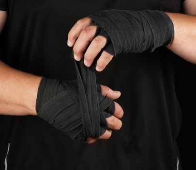 adult athlete stand in black clothes and wrap his hands in black textile elastic bandage before training, black background, muscular body