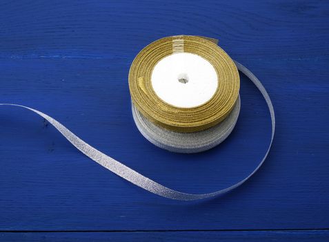 round bobbin with silver and golden ribbon on a blue wooden background, top view