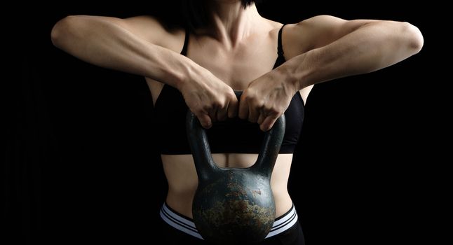 woman with a sports figure dressed in a black top holds a metal kettlebell in front of her, muscle tension, bodybuilding, low key